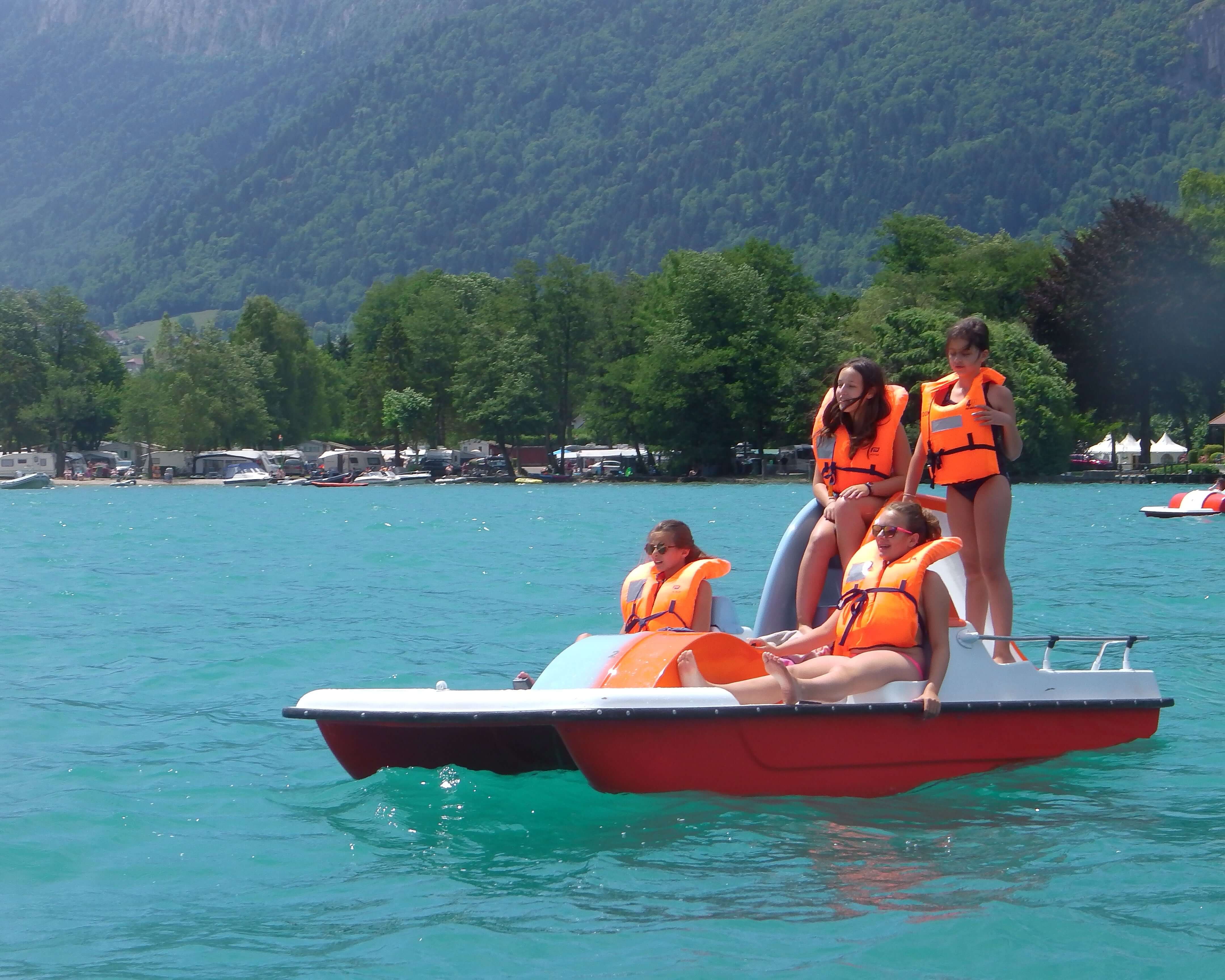 Teens in life jackets enjoying a pedal boat ride on a turquoise lake in the French Alps during summer camp.