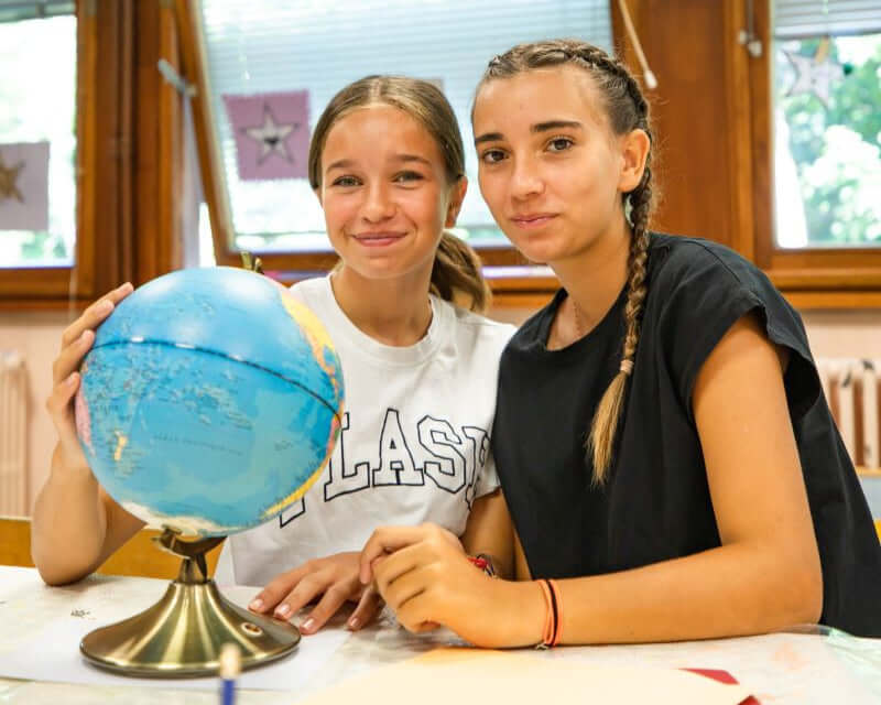 Two teenagers smile beside a globe, engaged in learning at a summer language camp in Megève, France.