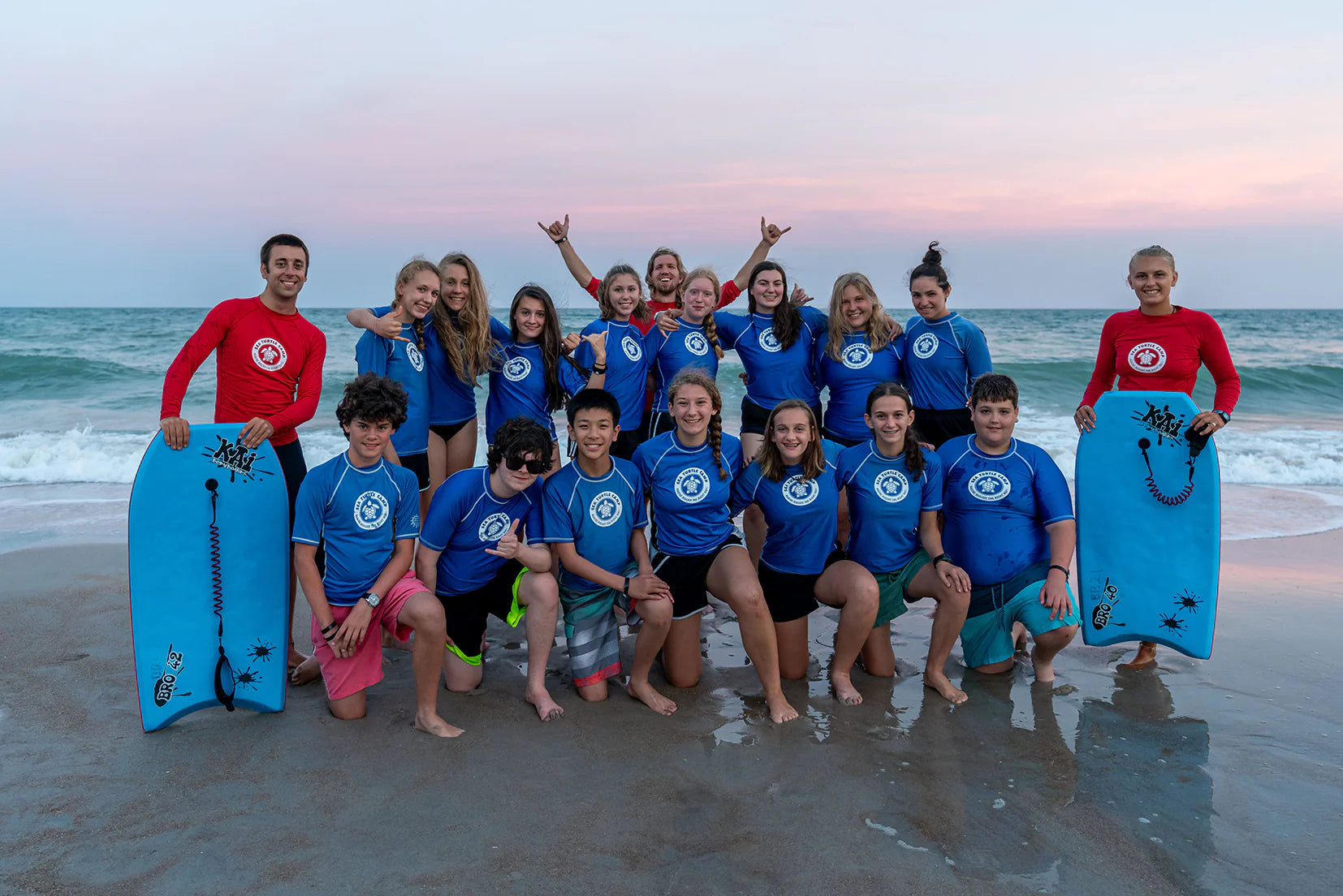 Group of teens in surf gear posing on the beach at sunset, enjoying summer programs and surf lessons.