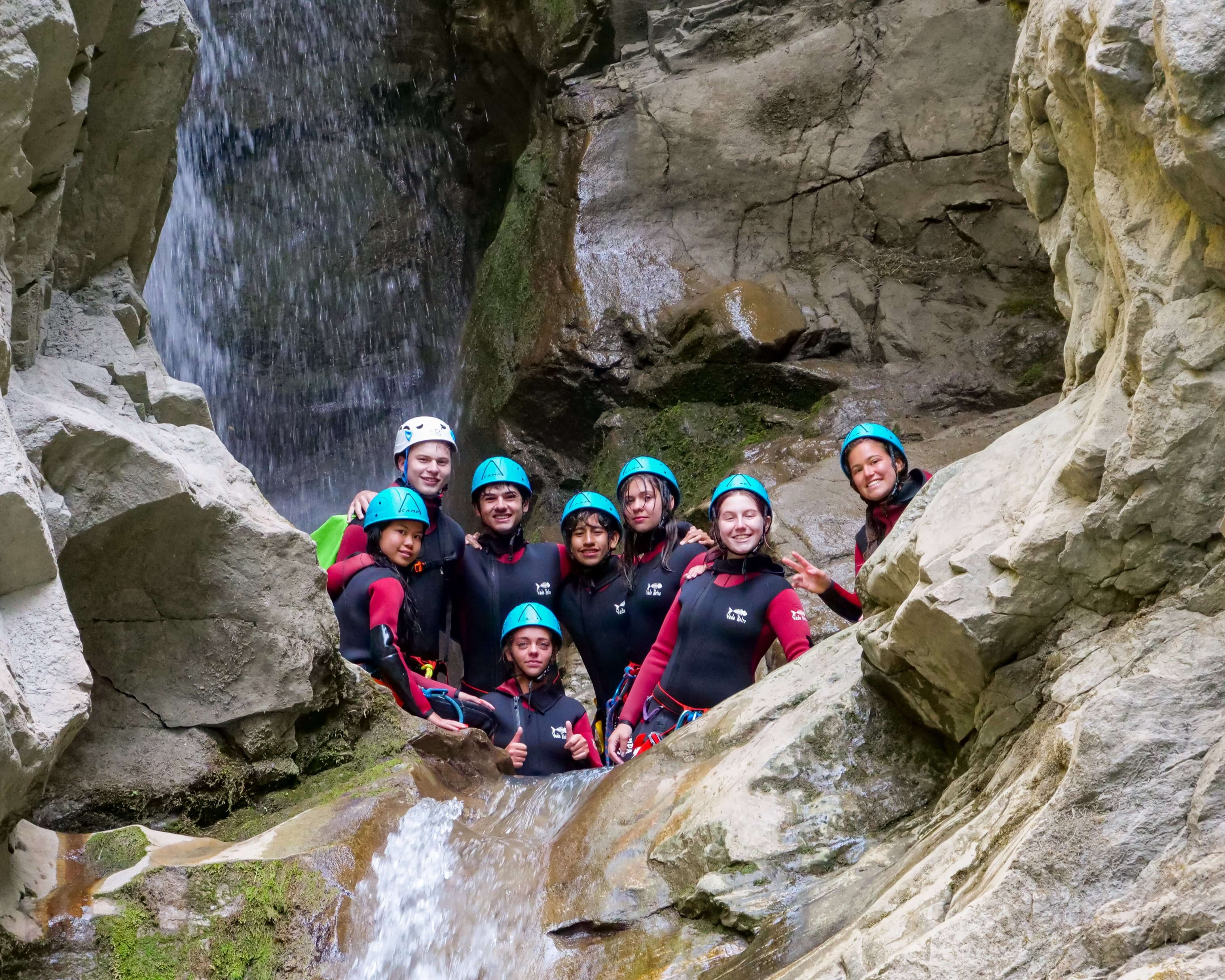 Group of teens in wetsuits smiling by a waterfall during a summer adventure activity in the French Alps.