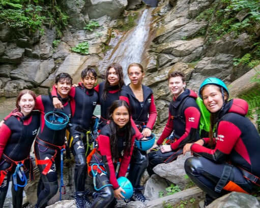 Group of teens in wetsuits enjoying canyoning adventure at a waterfall in Megève, France during summer camp.