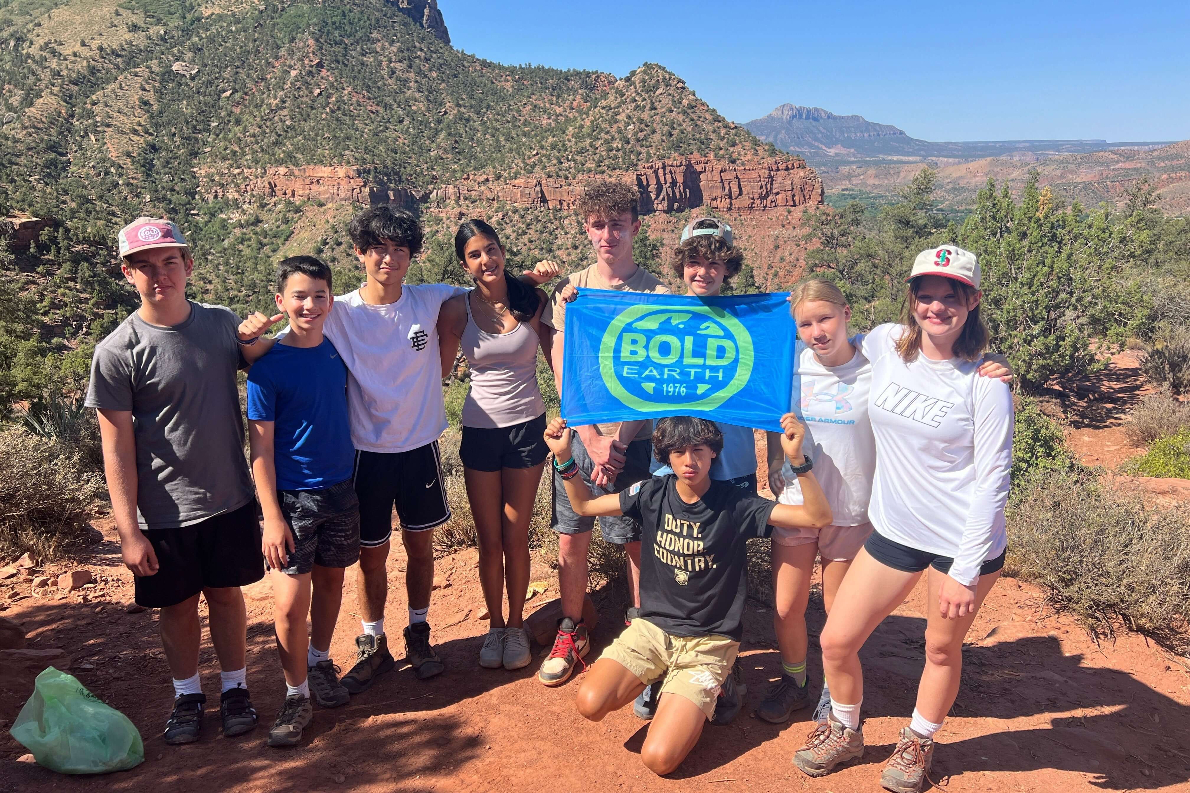 Group of teenagers holding a Bold Earth banner during a summer adventure trip in a scenic outdoor setting.