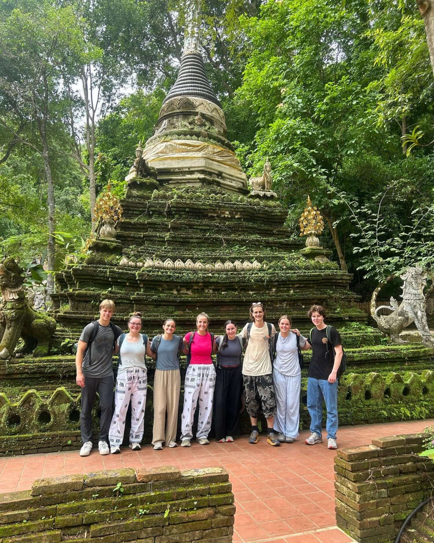 Group of teenagers in a lush forest, standing by a historic temple during their summer adventure trip.