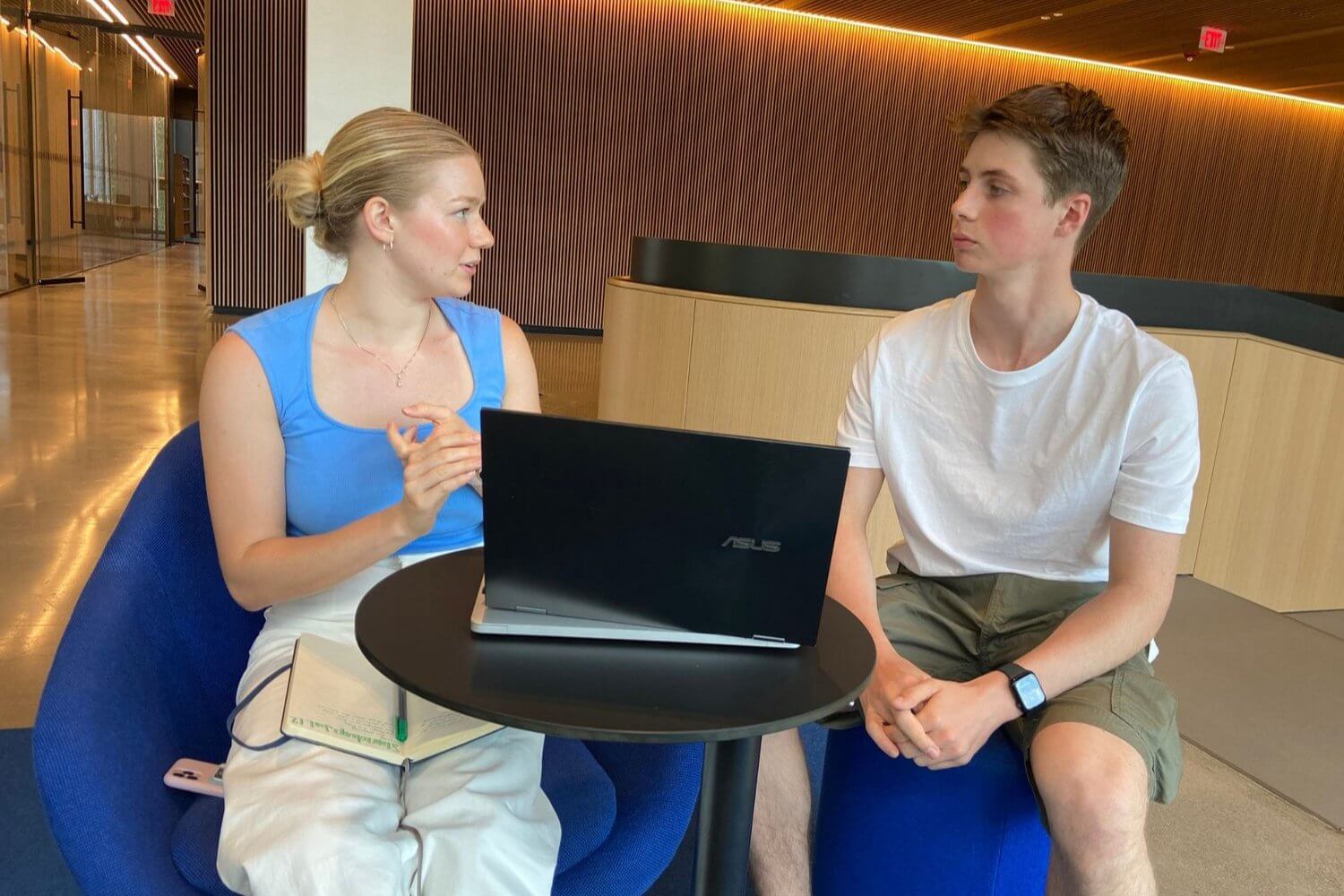 Two young students engage in discussion during a summer program, with a laptop on their table.