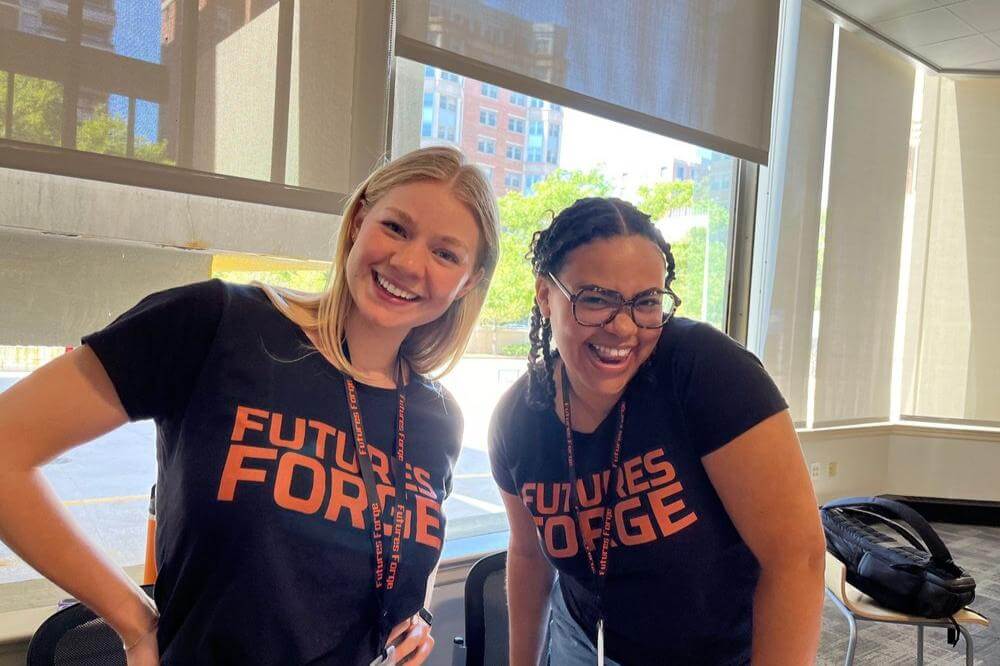 Two smiling women wearing Futures Forge shirts in a bright classroom, promoting the summer program for high school students.