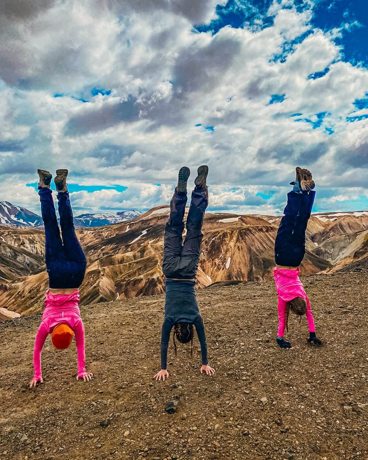 Three teenagers performing handstands on a mountain top, enjoying their summer adventure trip in a stunning, scenic landscape.