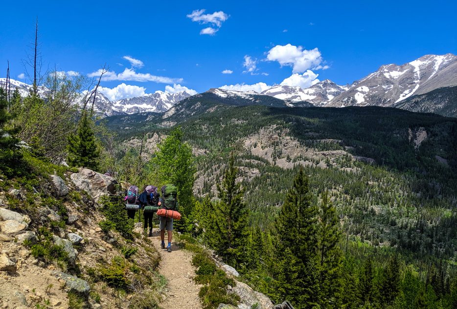 Hikers on a mountain trail in Colorado, surrounded by lush greenery and stunning snow-capped peaks. Perfect for summer programs.