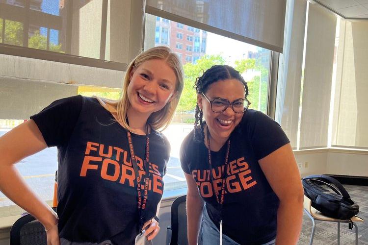 Two smiling young women wearing "Futures Forge" t-shirts in a bright room, promoting teamwork and skills development.