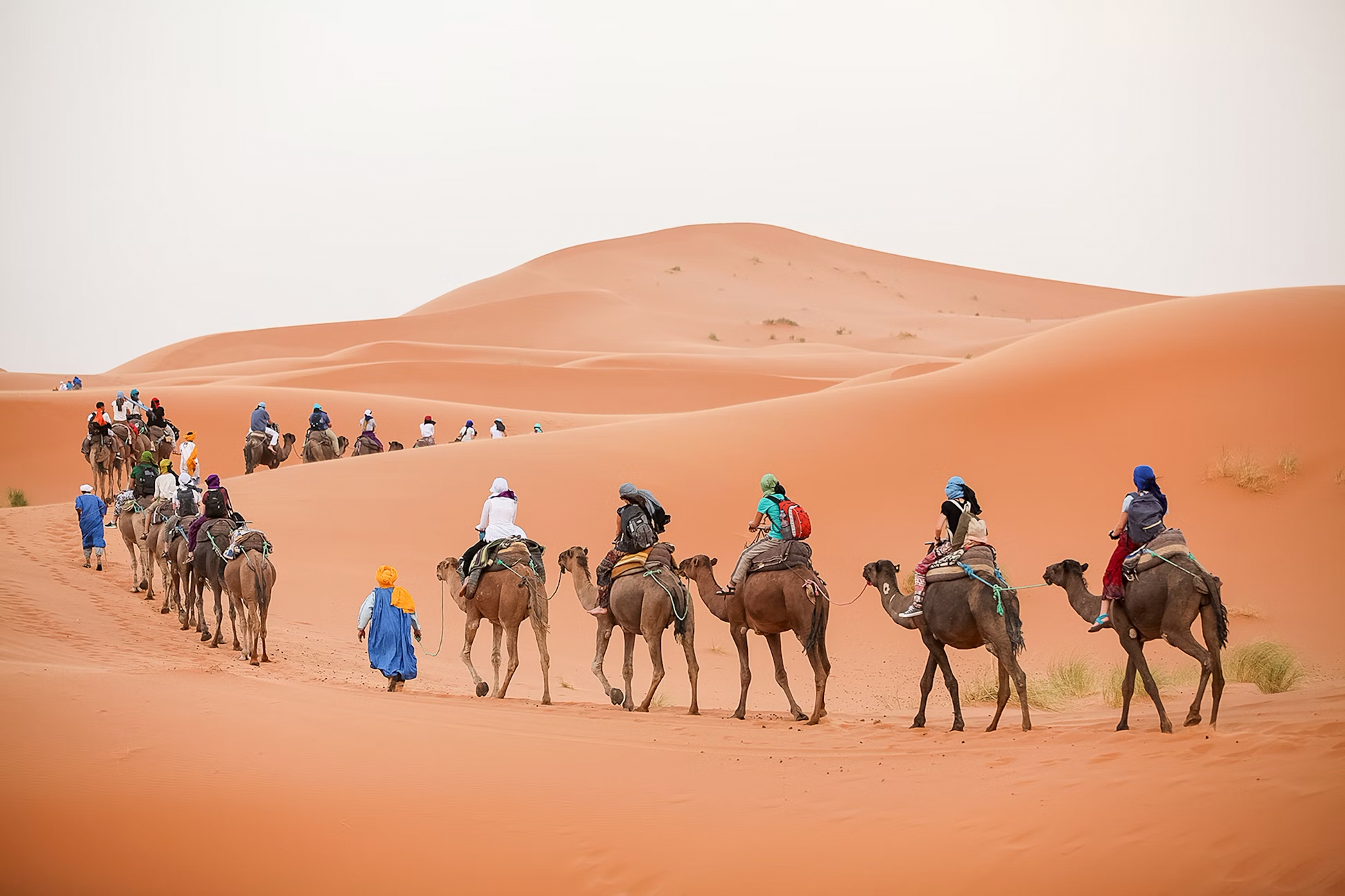 A caravan of camels and travelers walking through vast orange sand dunes in a desert landscape.