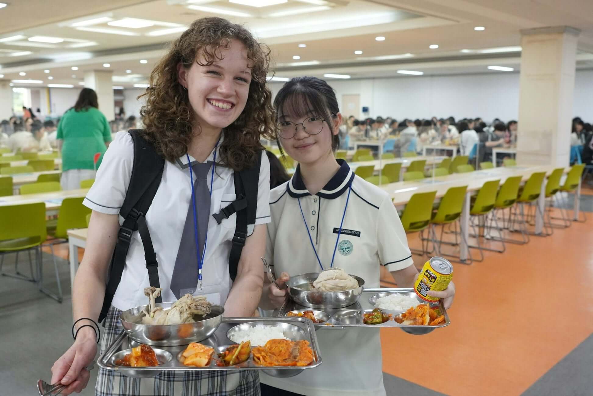 Two teens smiling while holding trays of traditional South Korean food in a school cafeteria setting.