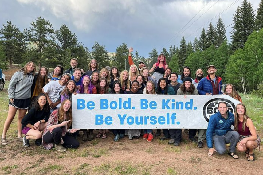 Group of diverse young people holding a banner that says "Be Bold. Be Kind. Be Yourself." in a natural outdoor setting.