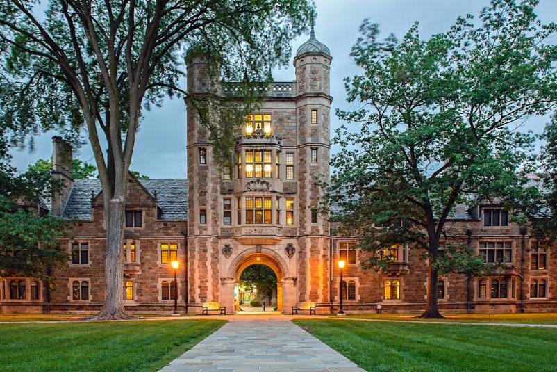 Historic college building with archway and trees in foreground, illuminated at dusk.