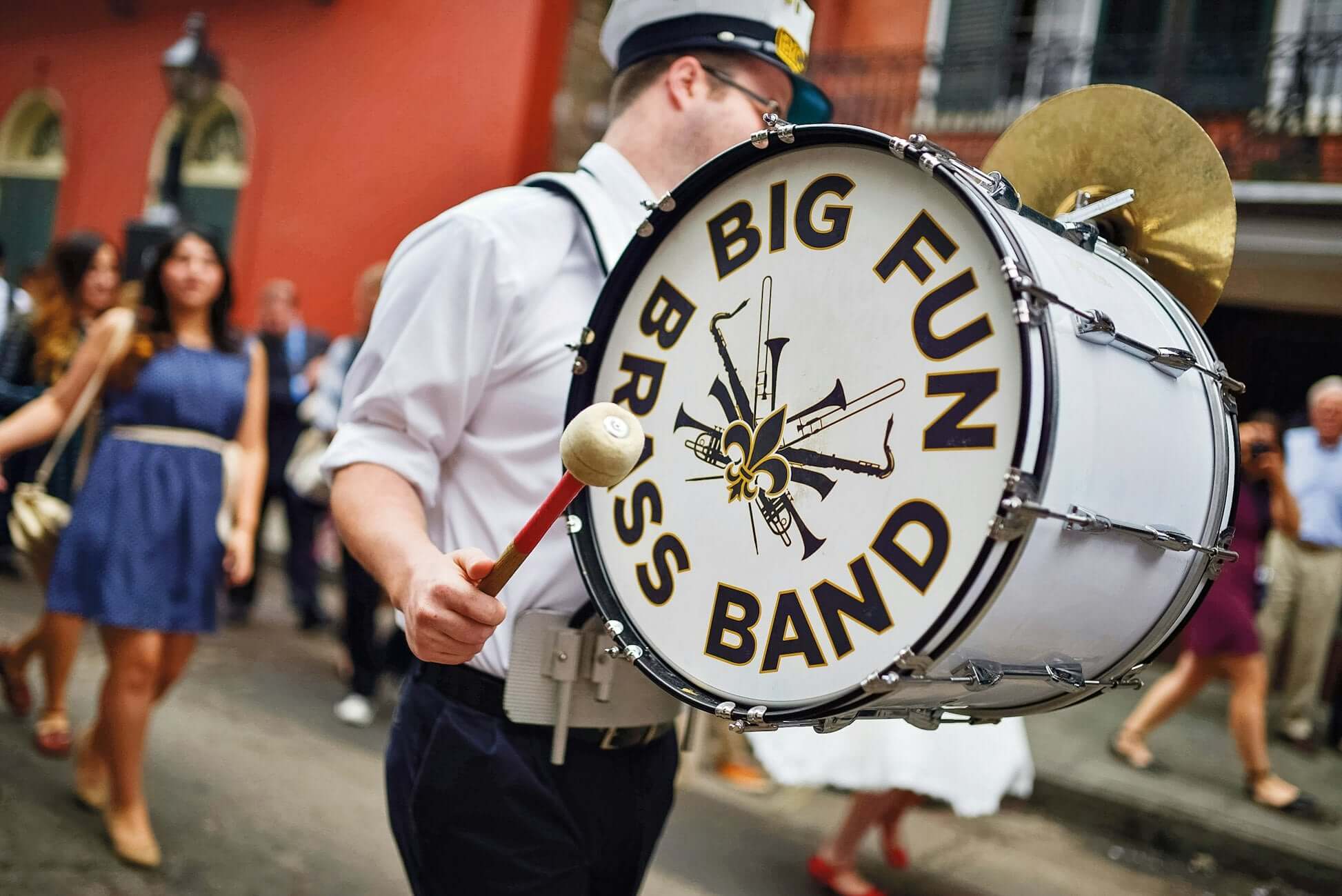 A musician in a white uniform plays a large drum marked "Big Fun Brass Band" in a lively New Orleans street parade.