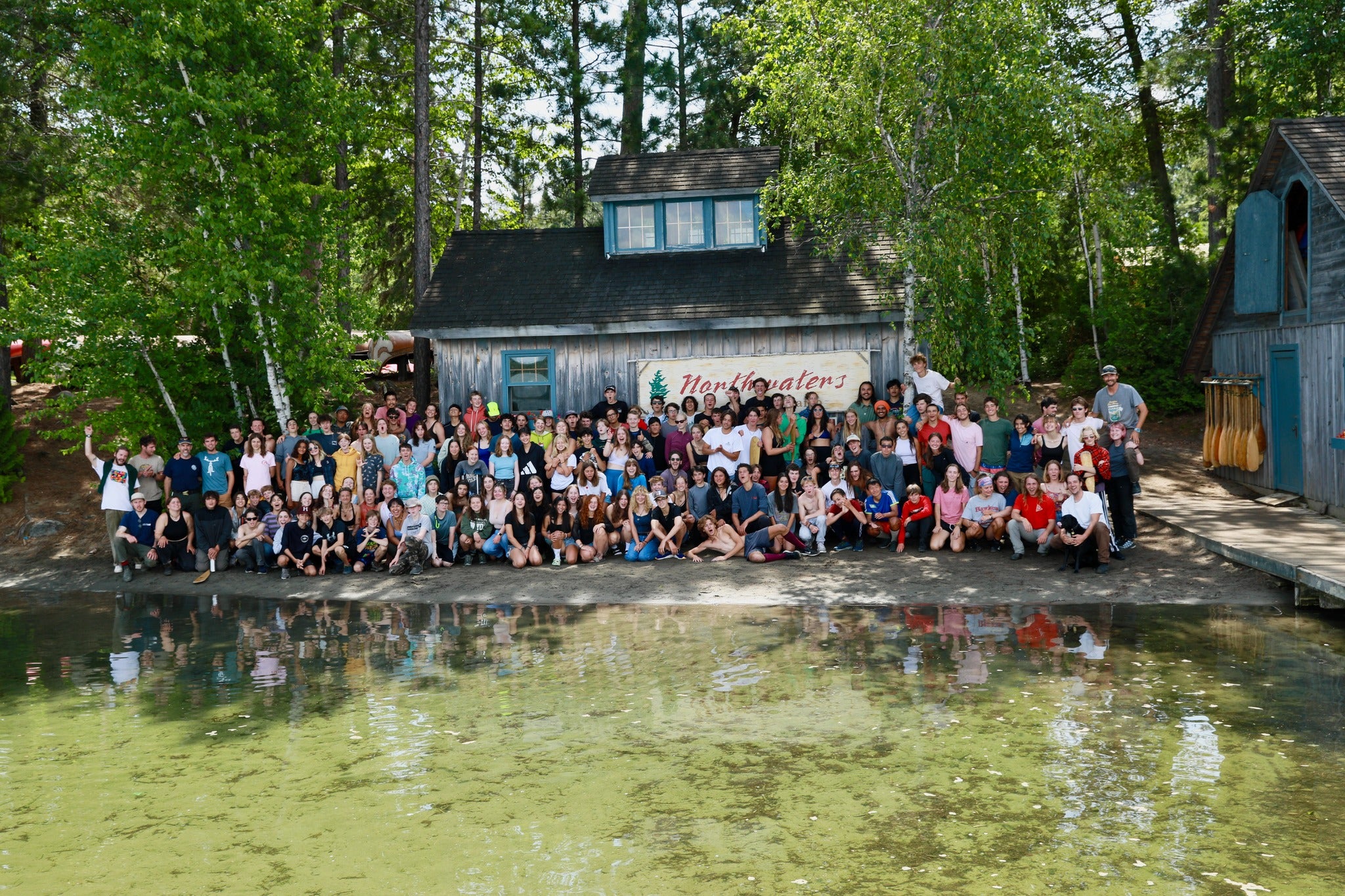 Large group of summer campers by the water in front of a cabin, showcasing a vibrant summer programs experience.