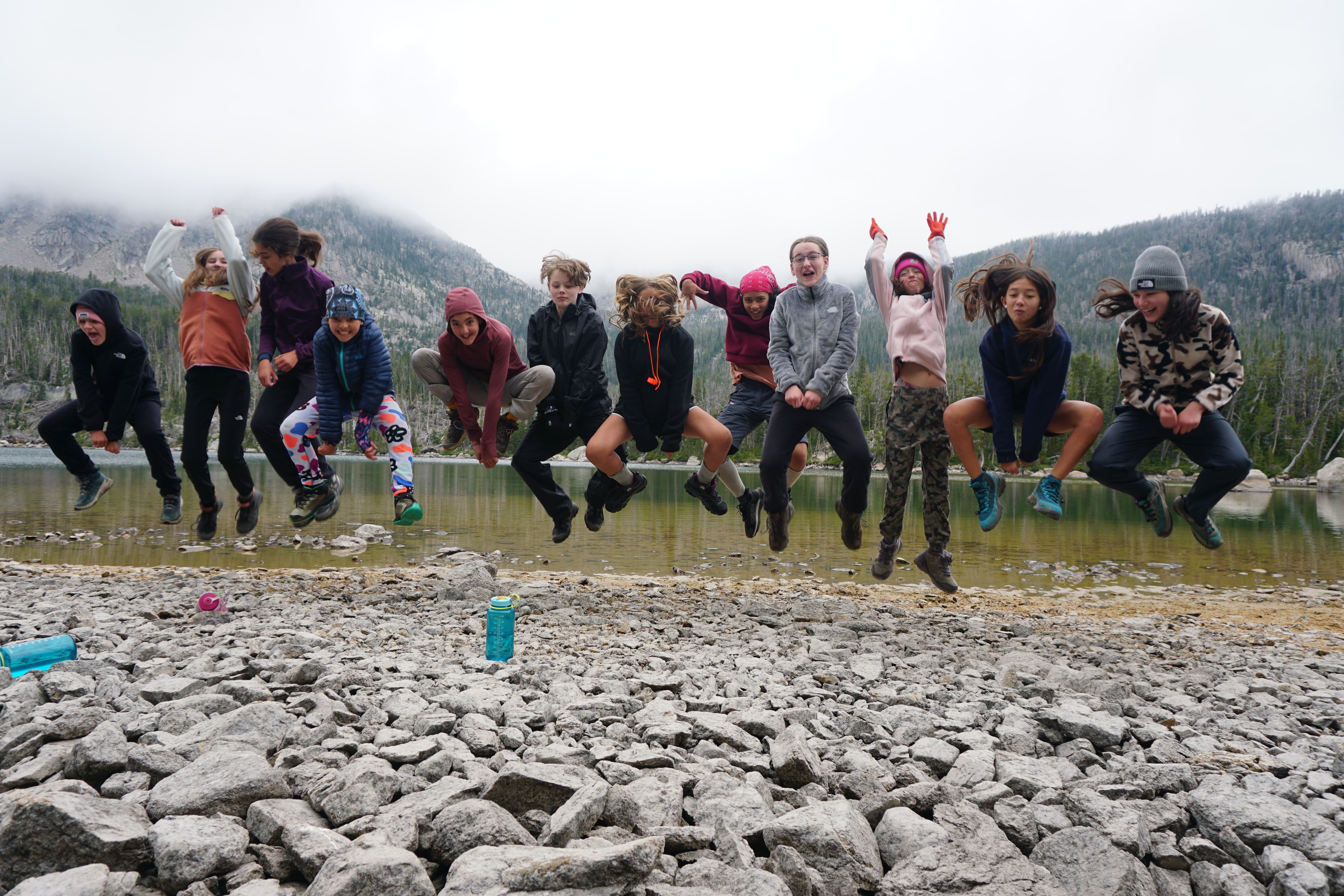 Group of teens joyfully jumping on rocky shore during Alpengirl Camp summer program, surrounded by nature.